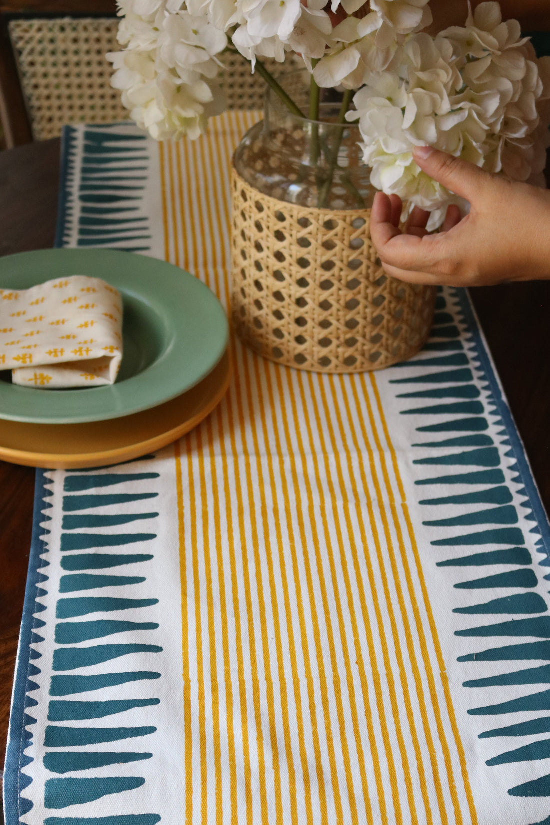 a cotton table runner with mustard stripes in the middle and a teal blue triangle border on both sides. the photo shows a vase of white flowers and a plate with a cotton napkin placed on it 